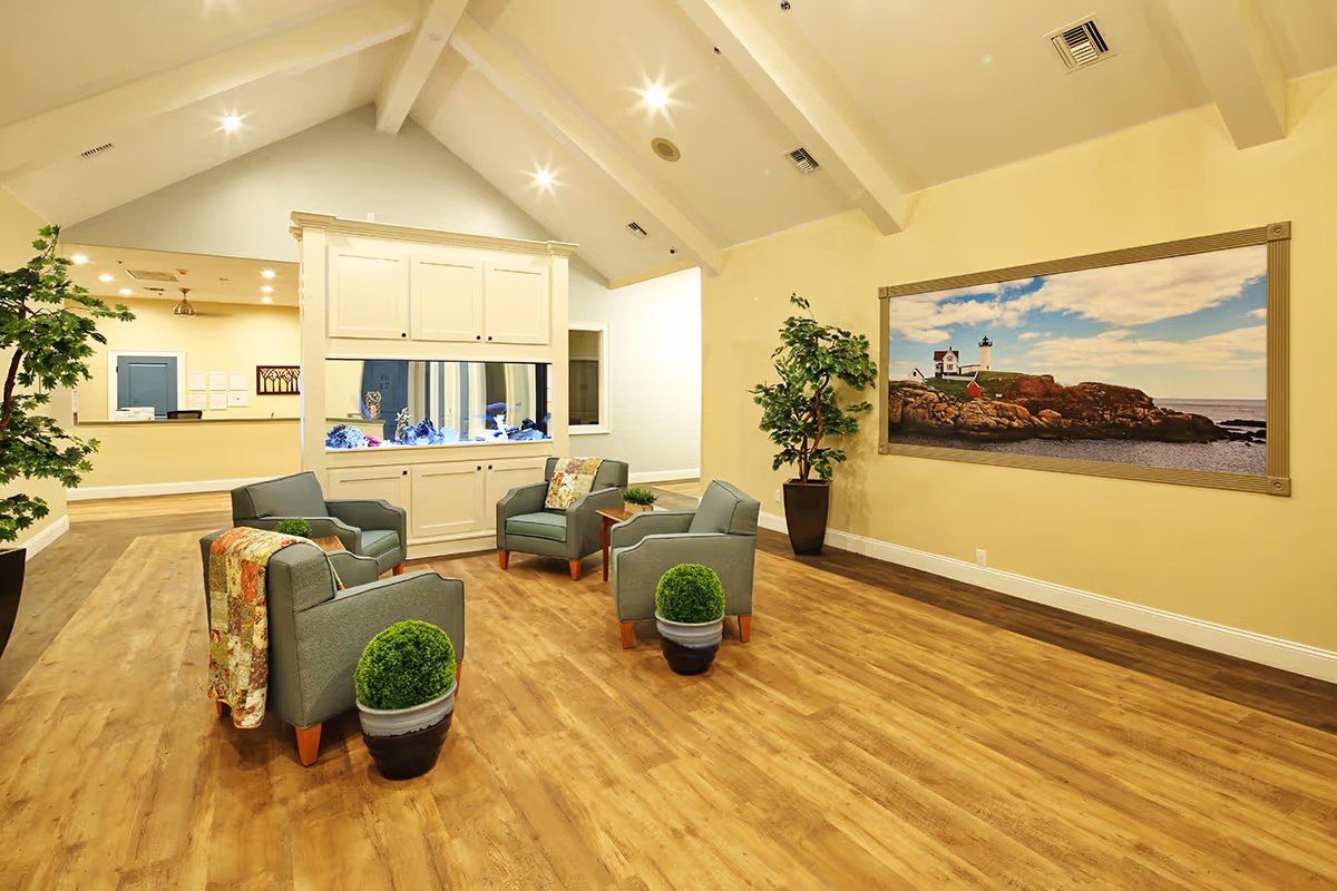 A spacious and well-lit common area in an assisted living facility featuring four gray armchairs arranged around a white cabinet with a built-in aquarium. The room has wooden flooring, potted plants, and a large framed picture of a lighthouse on a rocky coast hanging on the wall.