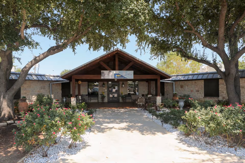 Entrance of Lake Village Nursing & Rehabilitation Center with a covered porch, surrounded by trees and landscaped bushes with pink flowers on either side of the walkway.