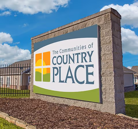 Large stone entrance sign reading "The Communities of Country Place" in front of residential buildings under a blue sky.