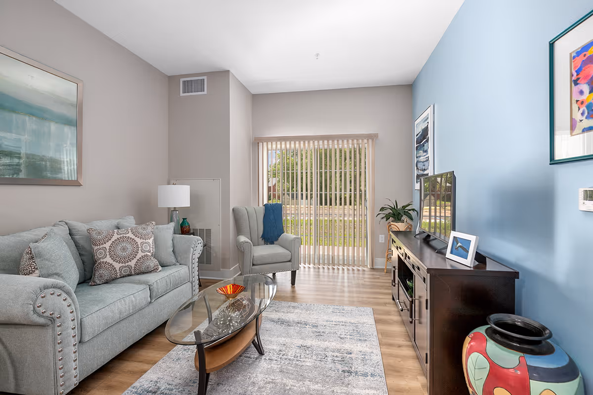A bright living room with a light blue sofa adorned with patterned cushions, a matching armchair with a blue throw, a glass coffee table on a patterned rug, a dark wooden TV stand with a flat-screen TV, framed artwork on the walls, a large window with vertical blinds letting in natural light, and a colorful decorative vase in the corner.