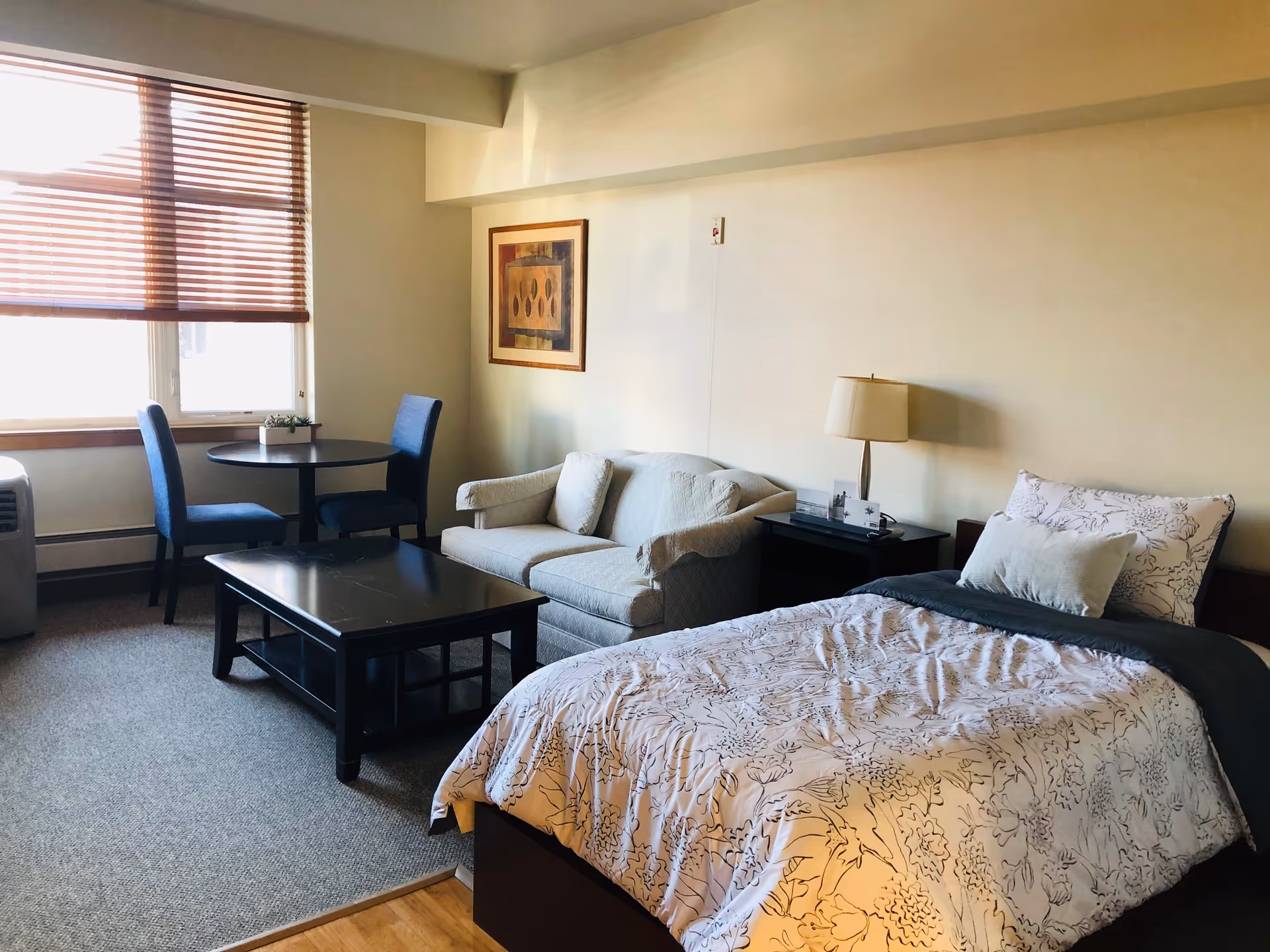 A well-lit senior living facility room featuring a single bed with floral bedding, a small beige loveseat, a dark wooden coffee table, a round dining table with two blue chairs, a lamp on a bedside table, and a window with wooden blinds.
