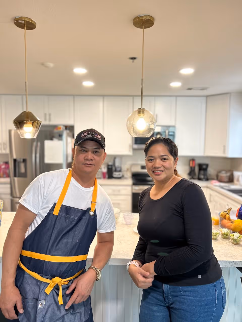 A man wearing a white t-shirt, dark apron with yellow straps, and a black cap stands next to a woman in a black long-sleeve shirt and blue jeans in a modern kitchen with white cabinets and a marble countertop. Two pendant lights hang from the ceiling above them.