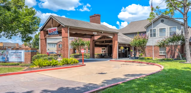 Exterior view of Lone Star Living senior living facility showing a covered entrance with a 'Now Leasing' sign, brick building, green lawn, trees, and a clear blue sky with clouds.
