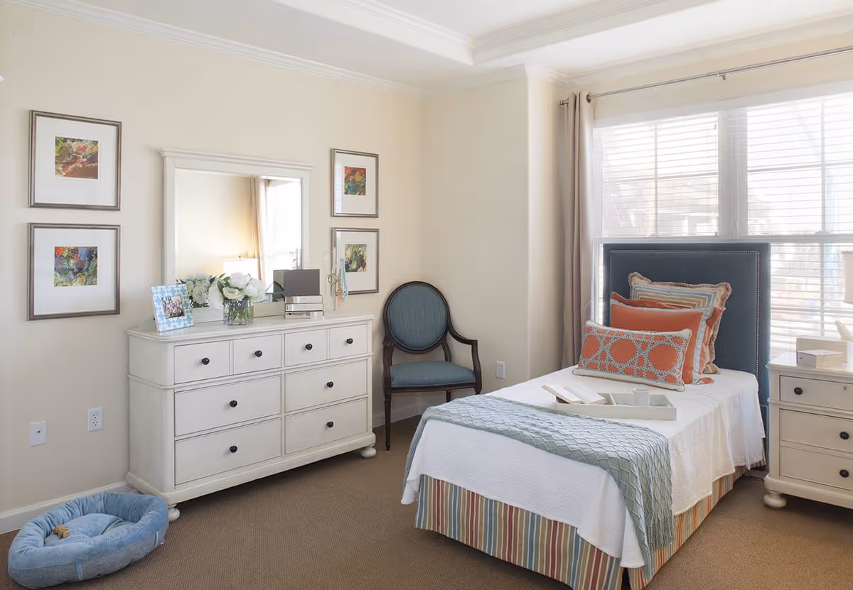 A bright and cozy bedroom with a single bed featuring a blue upholstered headboard and multiple decorative pillows. The bed is covered with a white quilt and a light blue throw blanket. Next to the bed is a white nightstand with drawers and a lamp. Across from the bed is a white dresser with multiple drawers, a mirror above it, and decorative items including a photo frame and a vase with flowers. A blue cushioned chair is placed in the corner near the window with white blinds and beige curtains. A blue pet bed is on the carpeted floor near the dresser. The walls are painted light beige and adorned with four framed colorful artworks.