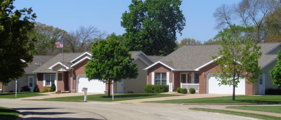 A row of single-story residential buildings with brick and siding exteriors, each with a garage and driveway. There are green trees and well-maintained lawns in front of the buildings under a clear blue sky.