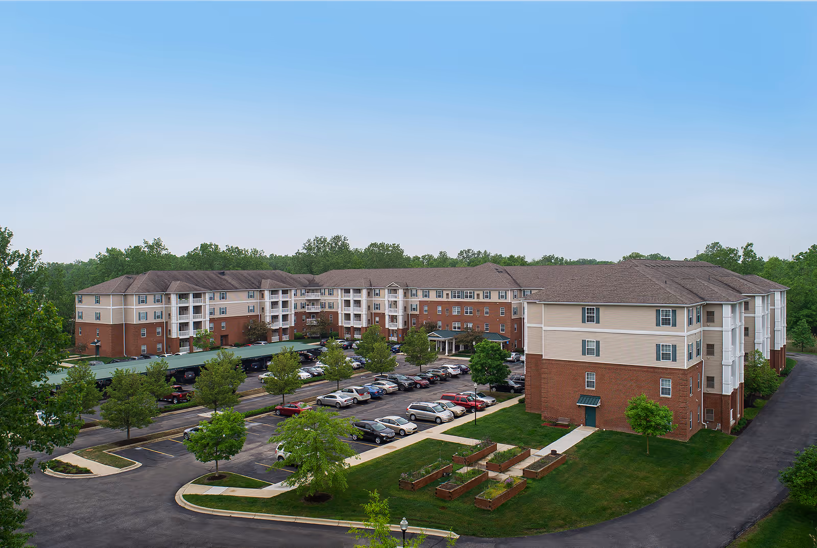 Exterior view of The Meadows of Brownstown senior living facility showing a large multi-story building with a parking lot in front, surrounded by green trees and landscaped garden beds under a clear blue sky.
