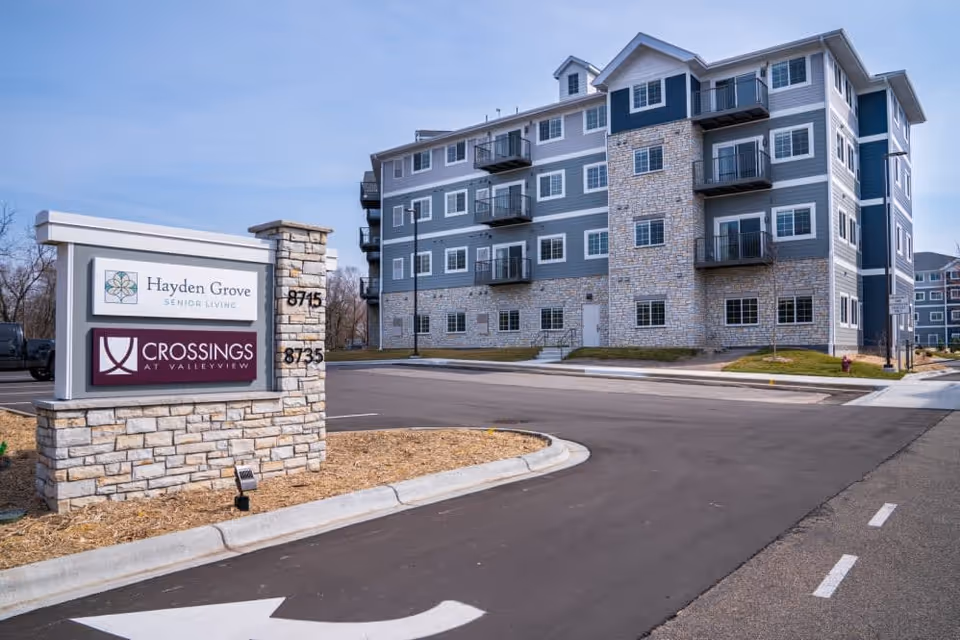 Exterior view of a multi-story senior living building with stone and siding facade and a roadside sign reading 'Hayden Grove' and 'Crossings at Valleyview'.