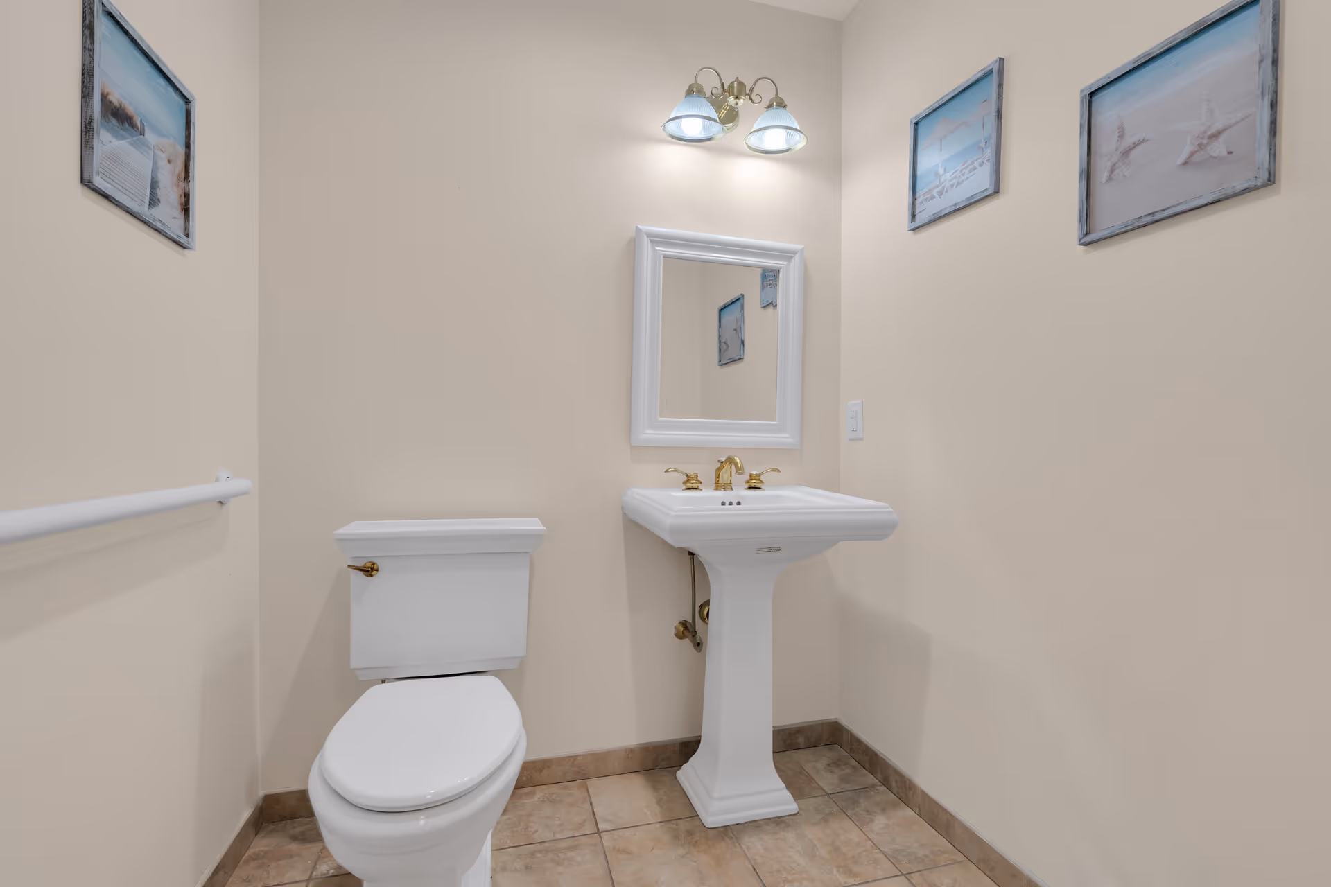 Bathroom with a toilet, pedestal sink, wall mirror and light, tile floor and framed pictures.