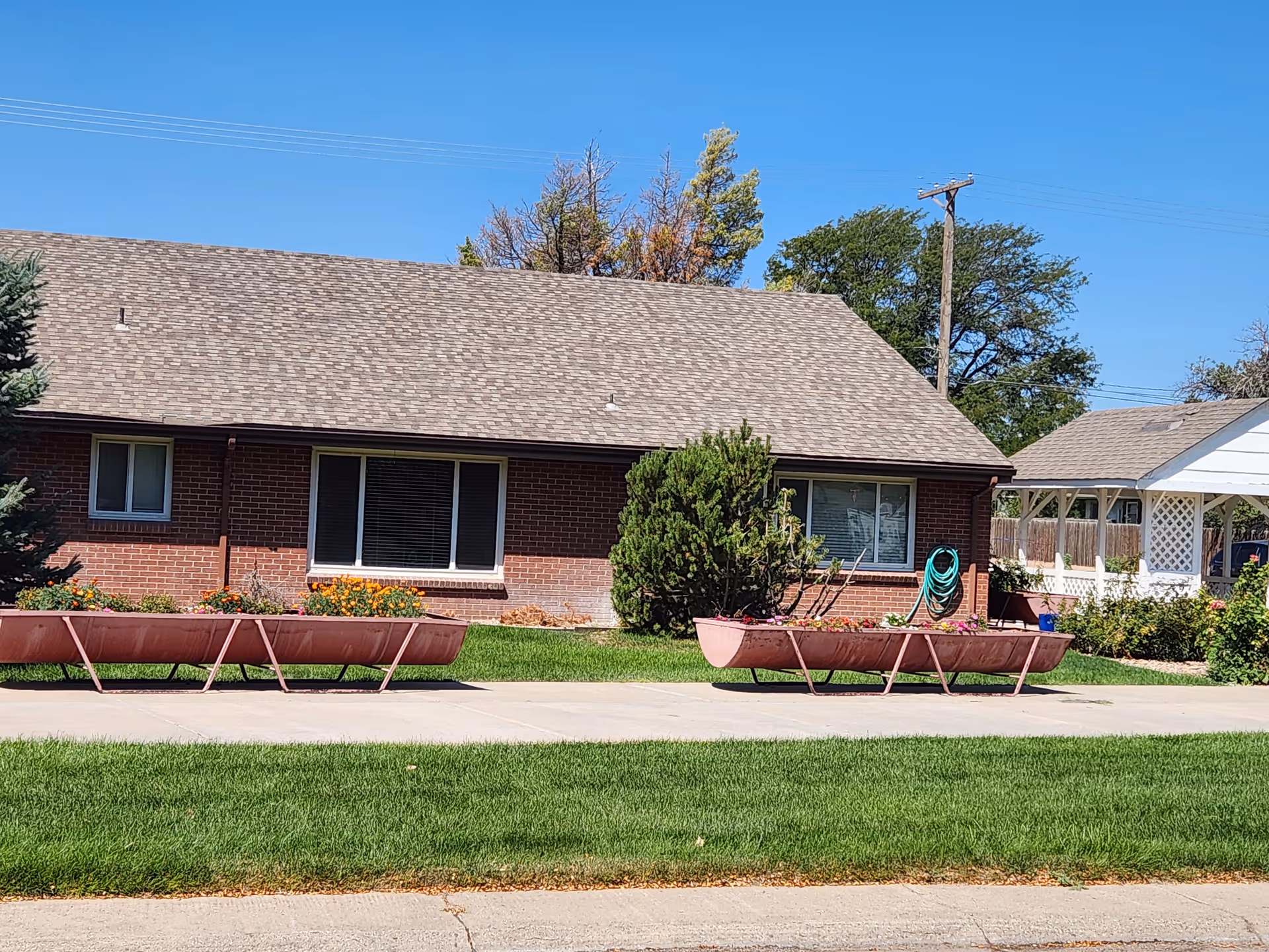 Front view of a one-story brick building with a green lawn, large planter troughs along the sidewalk, and a covered porch under a clear blue sky.