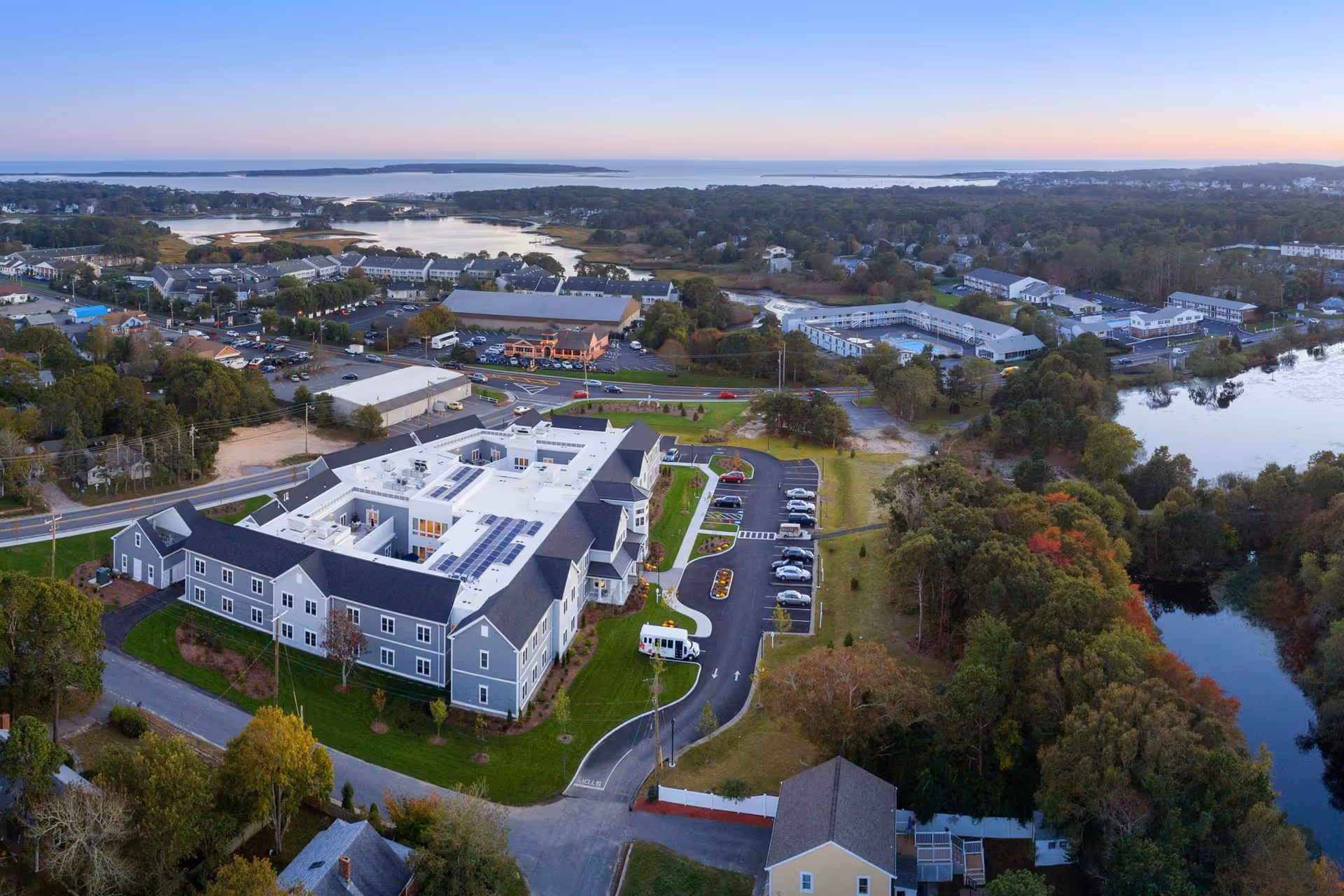 Aerial view of Maplewood at Mill Hill senior living facility surrounded by greenery, parking lots, nearby buildings, and water bodies under a clear sky at sunset.