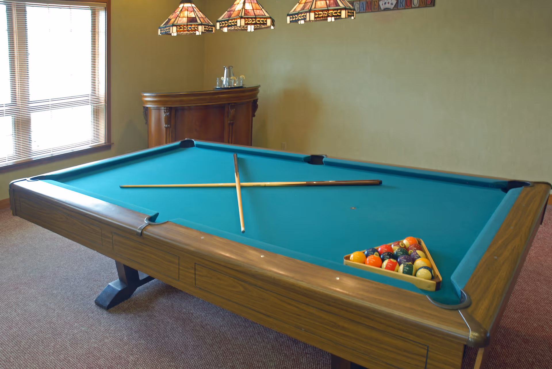 A billiards table with two cues crossed on the felt and a racked set of balls in a well-lit game room.