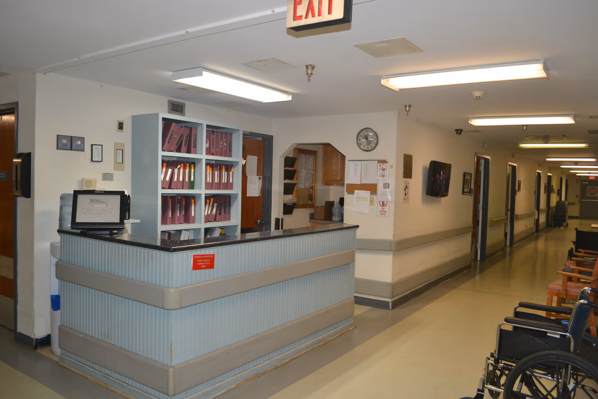 Reception desk area in a nursing home hallway with shelves holding binders behind the desk, an exit sign overhead, a clock on the wall, a television mounted on the wall, and several chairs and a wheelchair along the corridor.