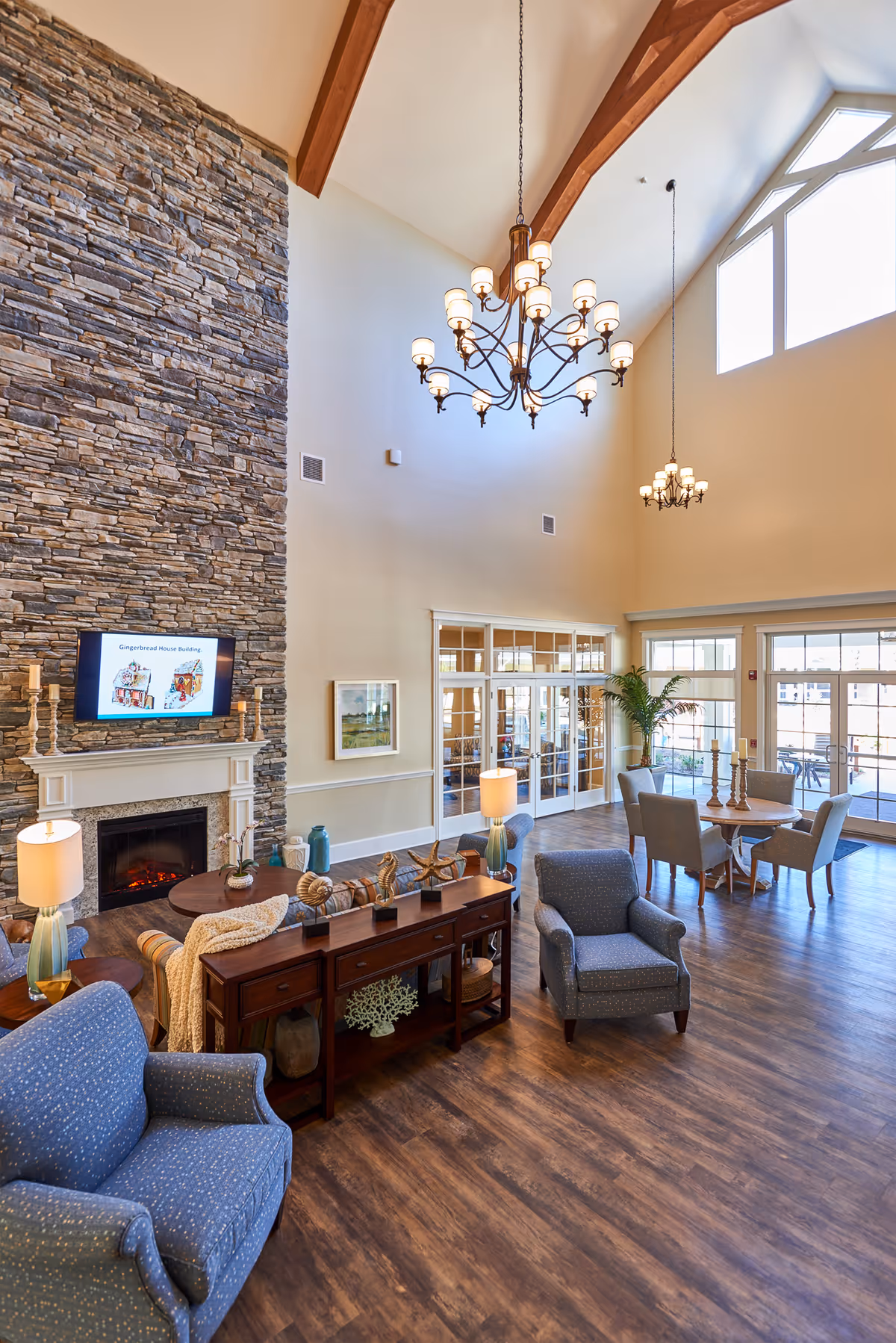 A spacious senior living facility common area with high vaulted ceilings and wooden beams. The room features a stone fireplace with a mounted TV above it, several comfortable blue armchairs, a wooden console table with decorative items, and a round dining table with chairs near large windows and glass doors that lead outside. Two elegant chandeliers hang from the ceiling, and the floor is covered with wood-style flooring.