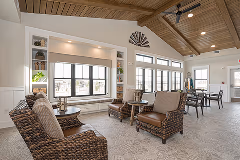 A spacious and bright living area with a high wooden ceiling and exposed beams. The room features large windows letting in natural light, a built-in window seat with cushions, and wicker armchairs with cushions arranged around small round tables. In the background, there is a dining table with chairs and a coat rack near the door.
