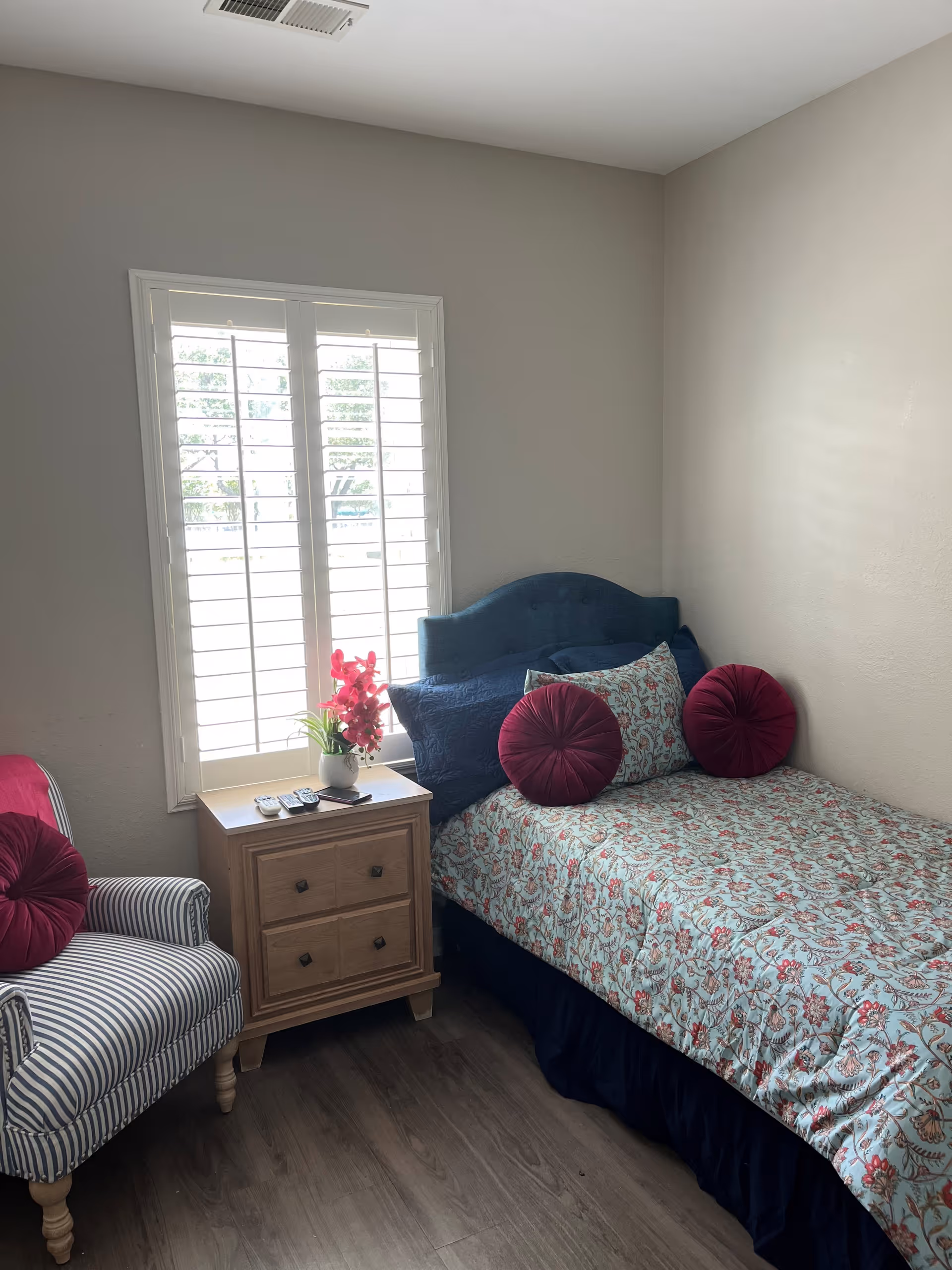 Small bedroom with a single bed featuring a patterned bedspread and round burgundy pillows, a striped armchair, and a nightstand by a window with shutters.