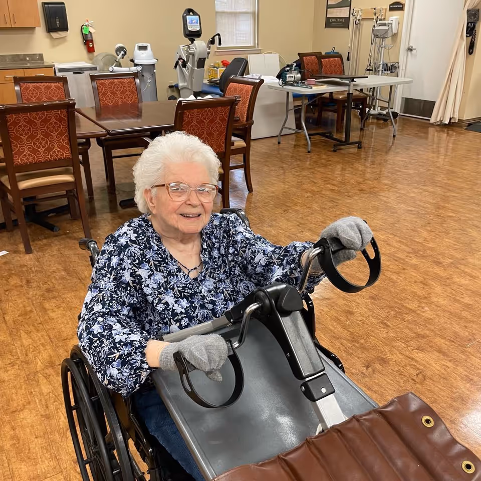 An elderly woman with white hair and glasses is sitting in a wheelchair, smiling and holding onto a piece of exercise equipment in a room with wooden floors and several tables and chairs in the background.