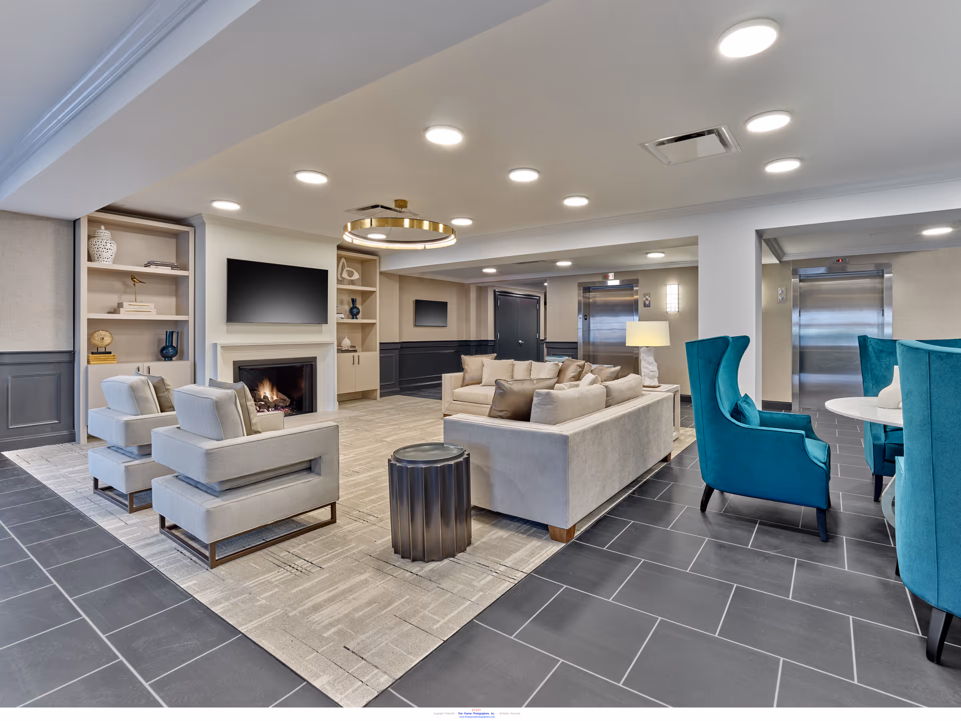 A modern senior living facility lounge area featuring a cozy seating arrangement with beige sofas and armchairs around a fireplace and wall-mounted TV. The room has built-in shelves with decorative items, a round table with teal high-back chairs, and two elevators in the background.