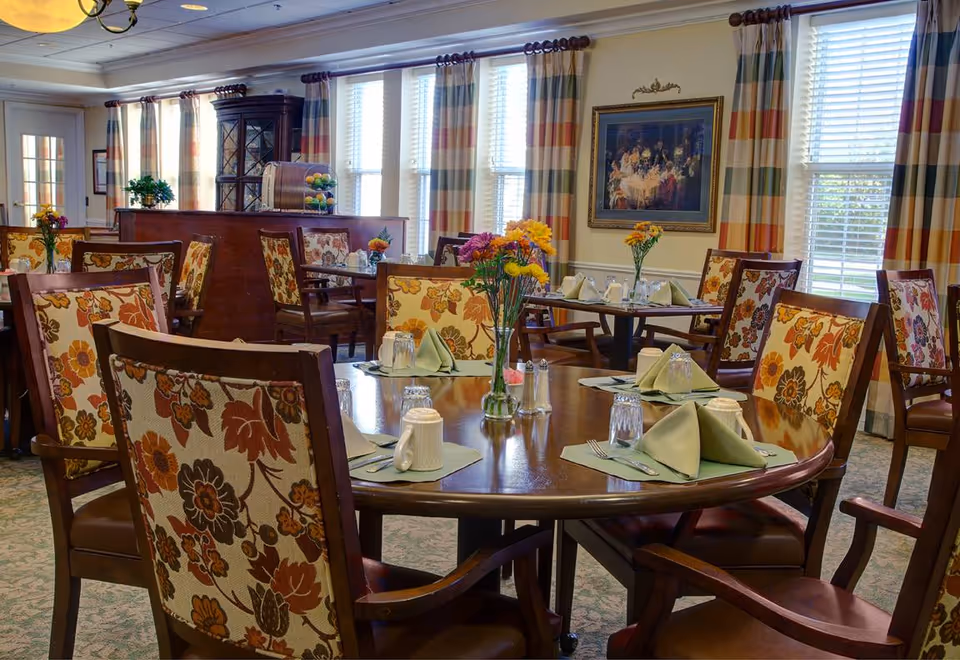 A dining room with round wooden tables set with green napkins, glassware, and silverware. The chairs have floral patterned upholstery. There are vases with colorful flowers on each table. The room has large windows with striped curtains and a framed painting on the wall.