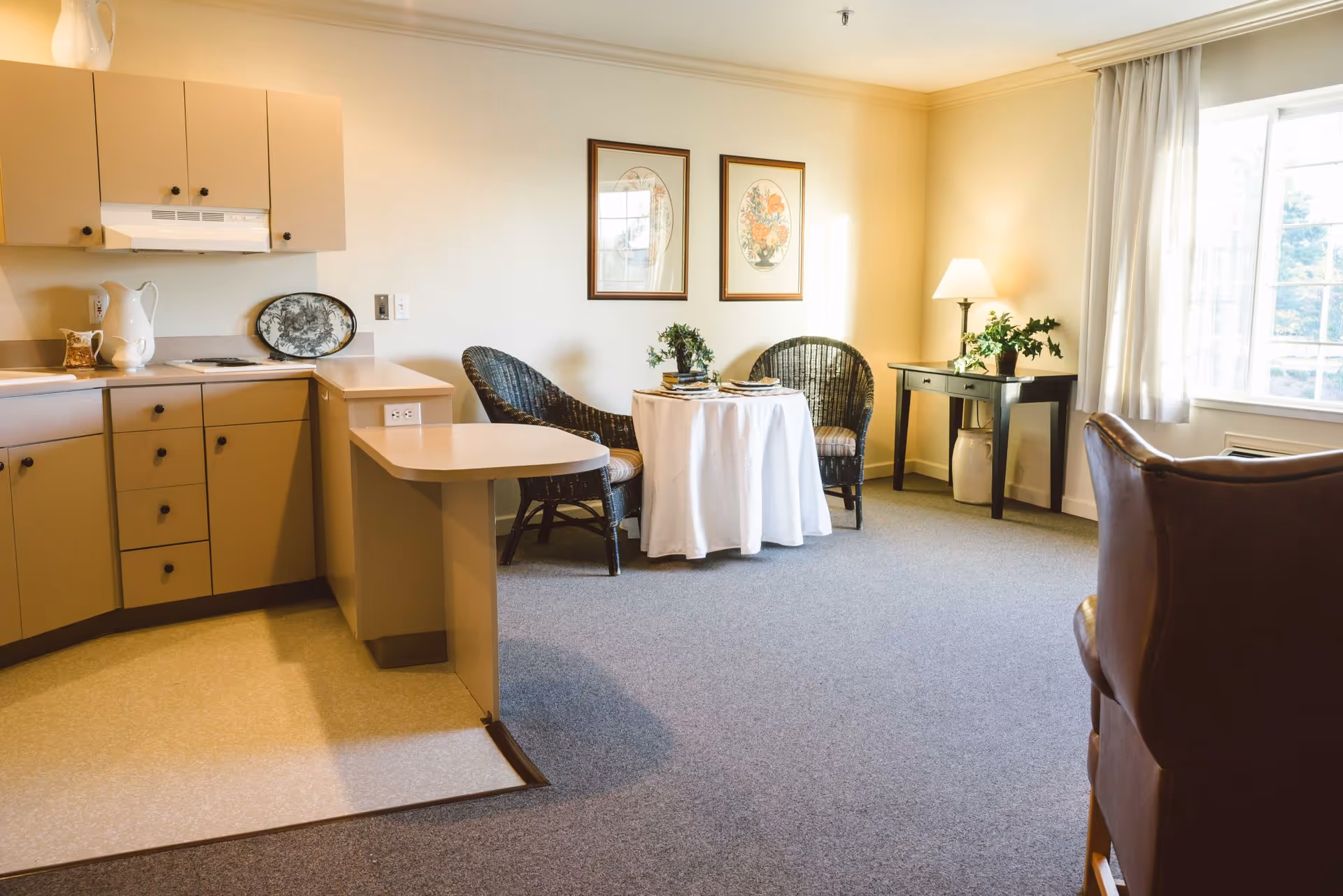 A cozy interior space featuring a small kitchen area with beige cabinets and a white countertop. Adjacent to the kitchen is a small round table covered with a white tablecloth, set with plates and surrounded by two wicker chairs. A window with white curtains allows natural light to brighten the room. There is also a dark wooden side table with a lamp and a plant, and a brown leather chair partially visible in the foreground.
