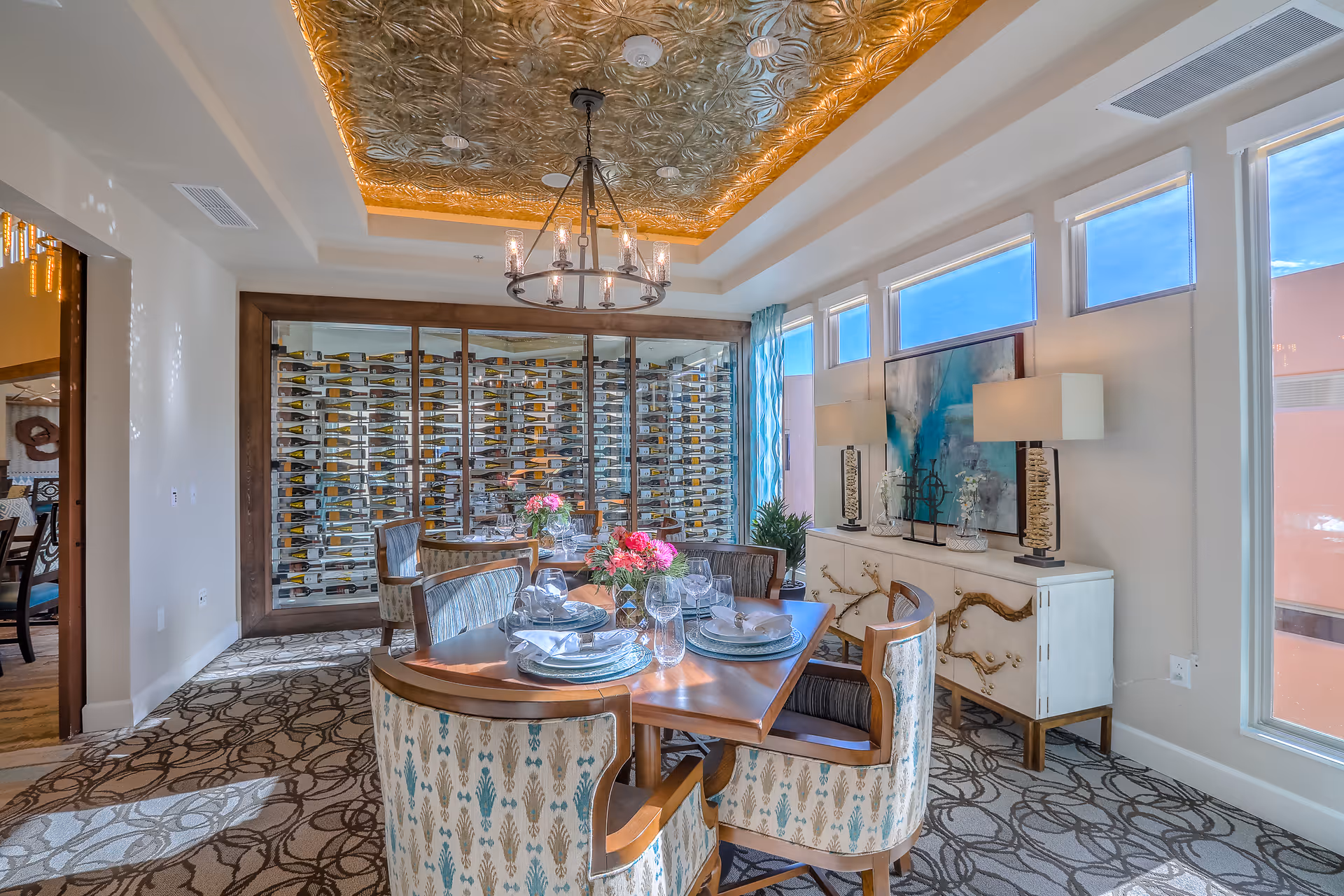 A well-lit dining room with a wooden dining table set for six people, featuring plates, glasses, and floral centerpieces. The room has patterned carpet flooring, a decorative gold ceiling with a chandelier, and a large glass-enclosed wine rack on one wall. There is a sideboard with two lamps and artwork above it, and several windows letting in natural light.