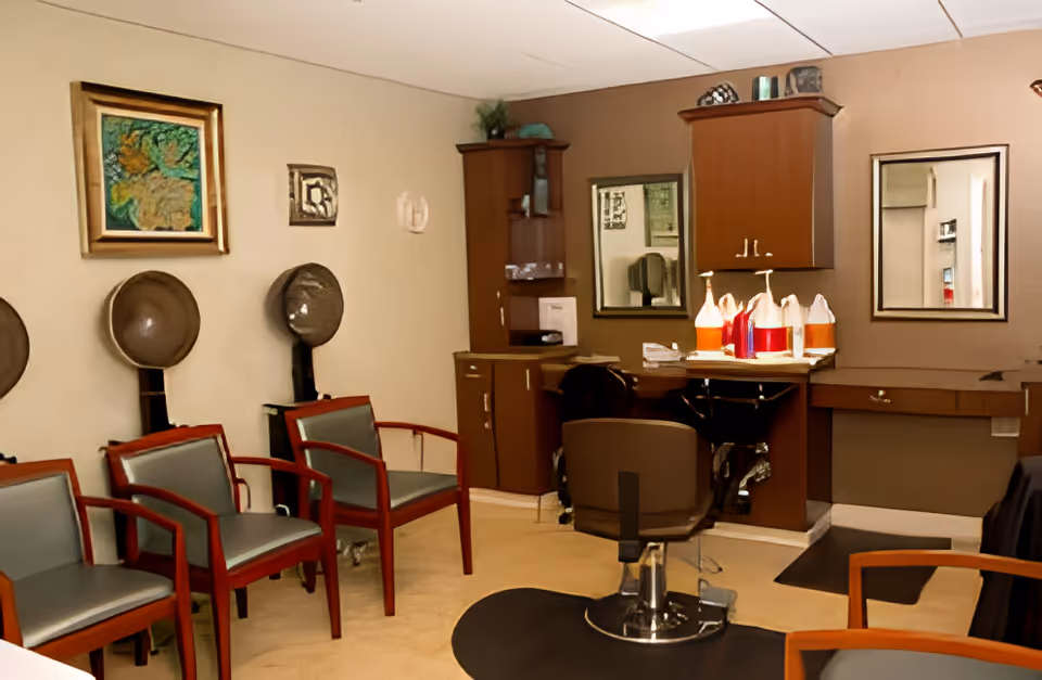 Interior view of a salon area with three vintage hair dryers mounted on the wall above chairs, a styling chair in front of a counter with hair care products, cabinets, and two mirrors on the wall.