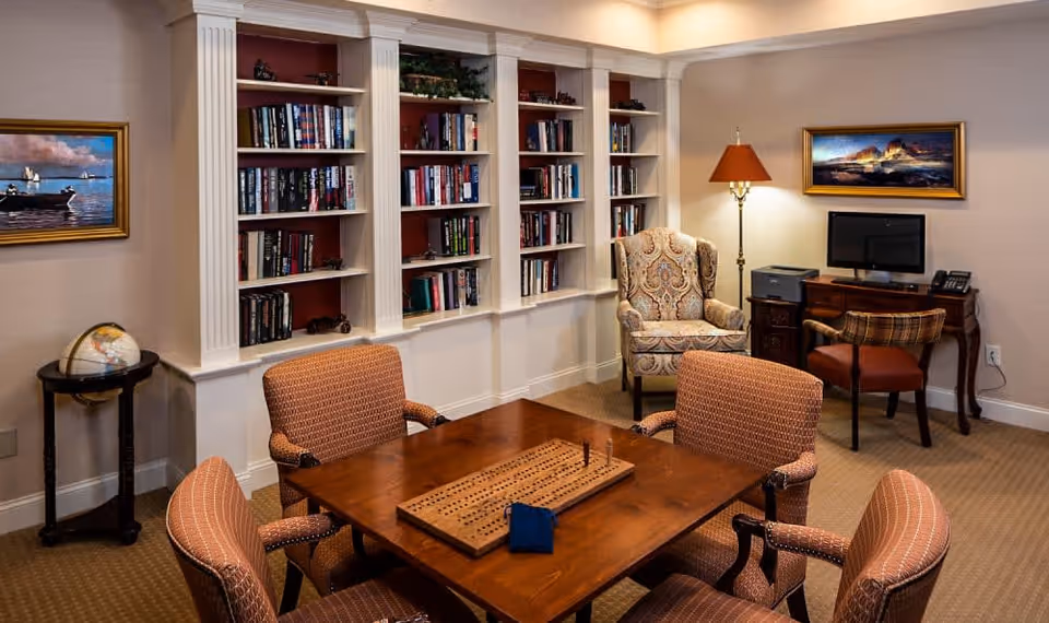 A cozy room with a wooden table and four upholstered chairs arranged around it. On the table is a cribbage board with pegs. Behind the table is a built-in bookshelf filled with books. To the right, there is a patterned armchair next to a floor lamp and a small wooden desk with a computer monitor, printer, and telephone. The walls are decorated with framed paintings, and a globe sits on a small round table in the corner.