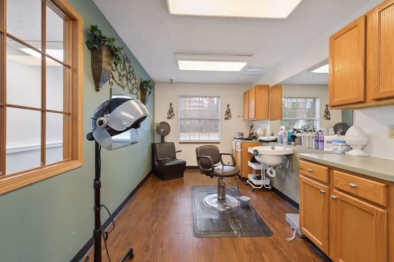 Interior of a hair salon area with a hair dryer on a stand, a salon chair in front of a sink, wooden cabinets, and a large mirror. The room has wooden flooring, a green accent wall with decorative wall hangings, and two windows letting in natural light.