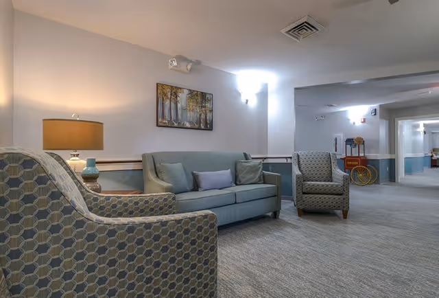 A cozy seating area in a senior living facility hallway featuring a light blue sofa with three cushions, two patterned armchairs, a side table with a lamp, and a framed painting of trees on the wall. The hallway extends into the background with soft lighting and a popcorn machine visible in the distance.