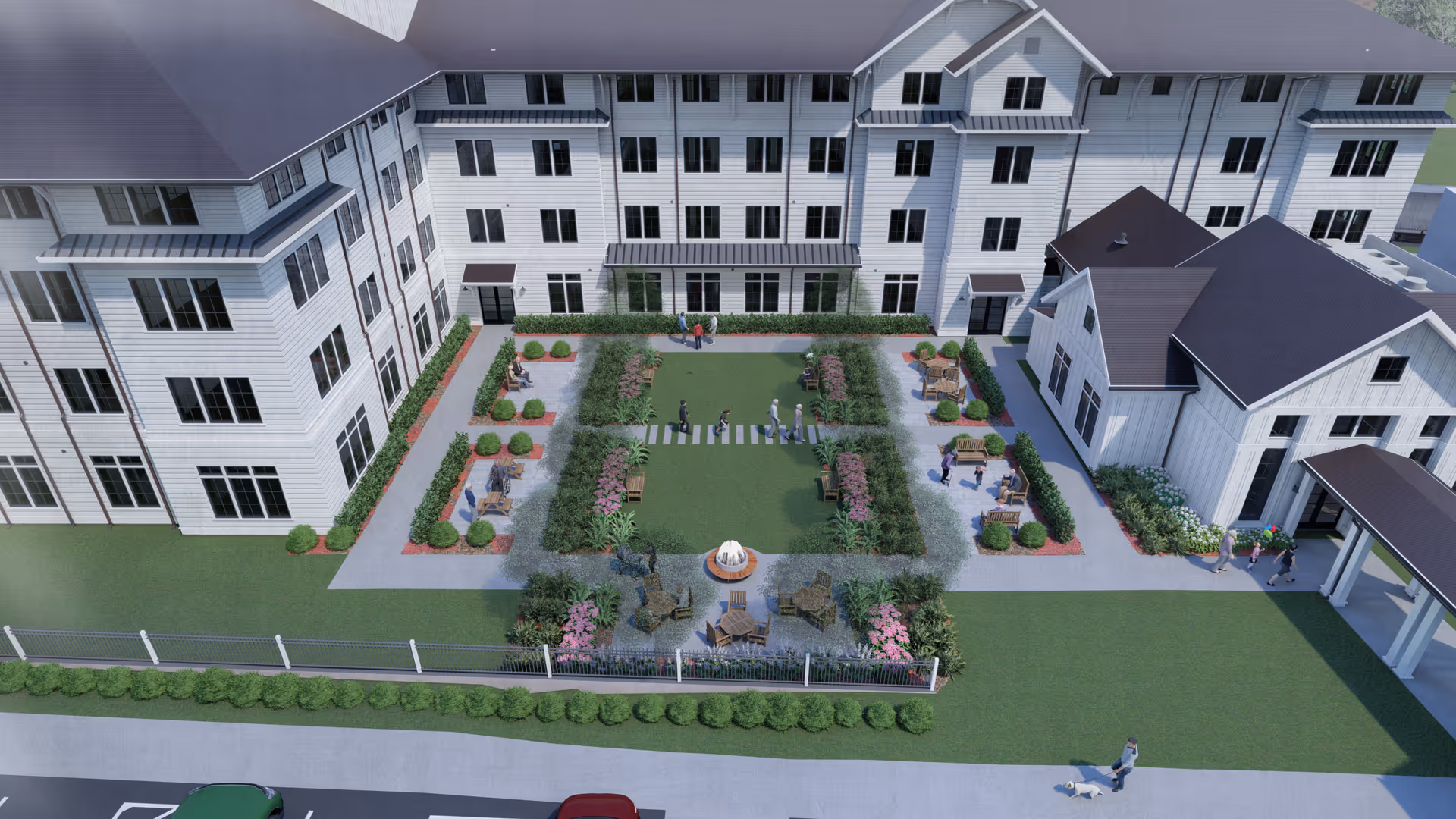 Aerial view of an outdoor courtyard area at Everlan of Clemson, featuring a central green lawn with people walking and sitting on benches. The courtyard is surrounded by a white multi-story building with numerous windows and landscaped garden beds with shrubs and flowers. There is a small fountain in the foreground and paved walkways connecting seating areas.