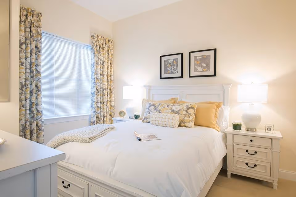 Bright, neatly made bedroom with a white bed, patterned curtains, two nightstands with lamps, and framed art above the headboard.