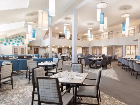 Spacious and well-lit dining room with multiple tables and chairs arranged neatly. The room features modern pendant lighting, a high ceiling with skylights, and decorative wall art with blue accents. The area has a mix of booth seating and individual chairs, creating a comfortable and inviting atmosphere.