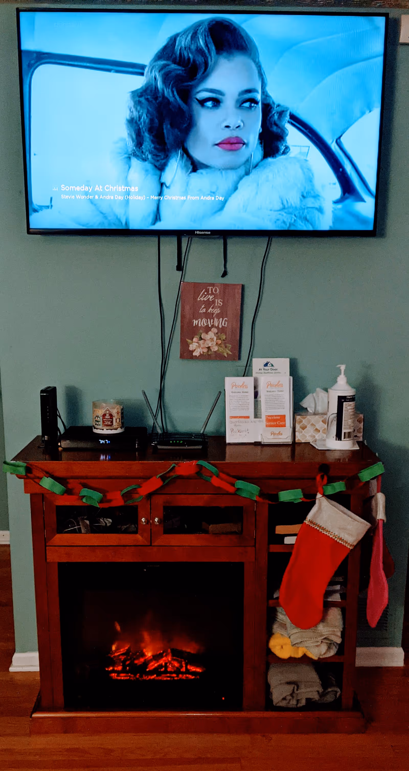Wall-mounted TV displaying a woman above a wooden media cabinet with an electric fireplace, holiday garland and stockings, and personal items on top.
