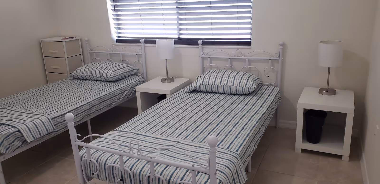 A simple bedroom with two single beds featuring white metal frames and striped bedding. Each bed has a pillow and a folded blanket at the foot. Between the beds is a white nightstand with a modern lamp, and another similar nightstand with a lamp is placed to the right of the right bed. There is a small drawer unit in the corner near the left bed. The room has tiled flooring and a window with horizontal blinds.