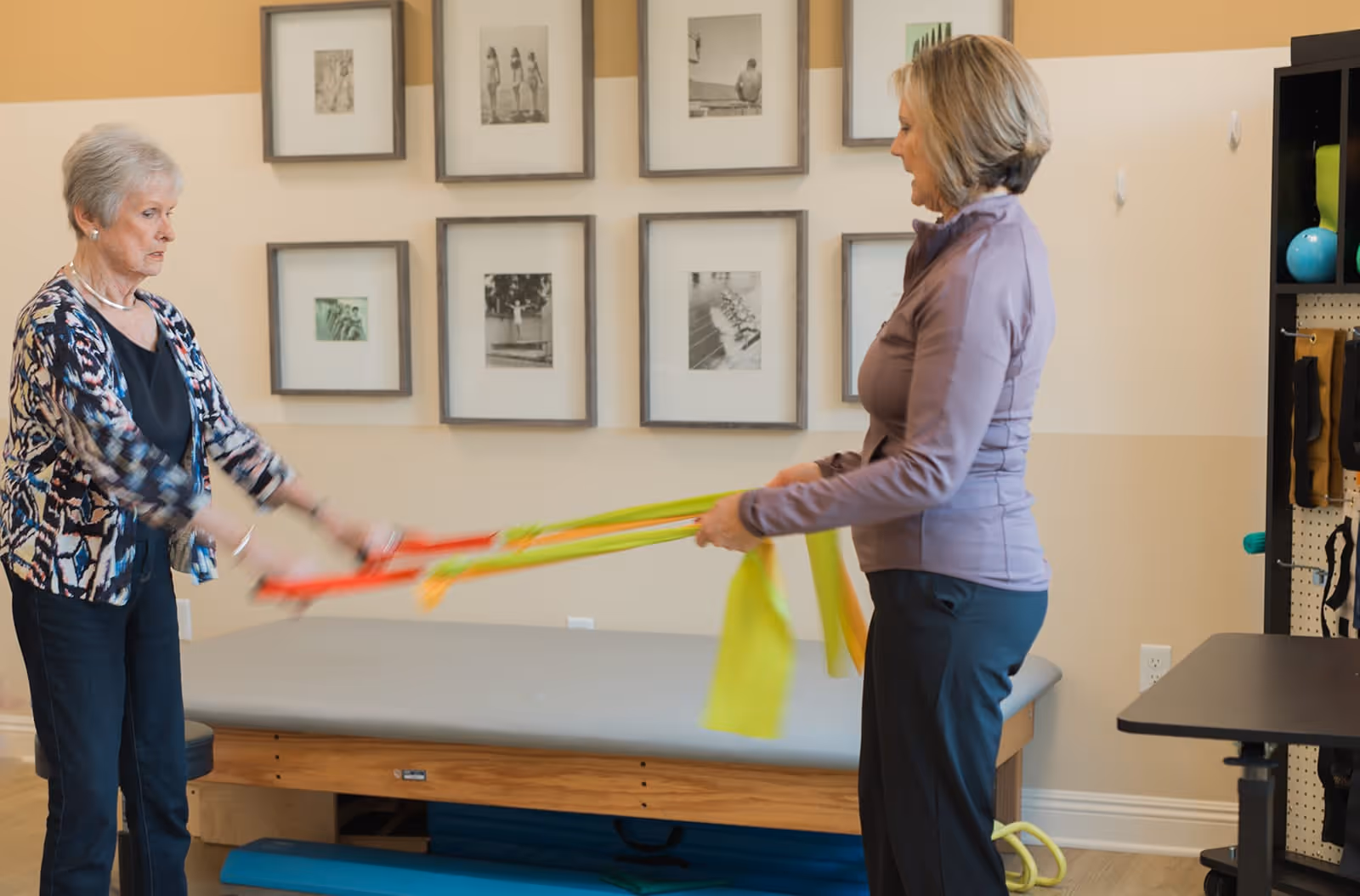 Two elderly women exercising with resistance bands in a room with framed black and white photos on the wall and a padded therapy table in the background.