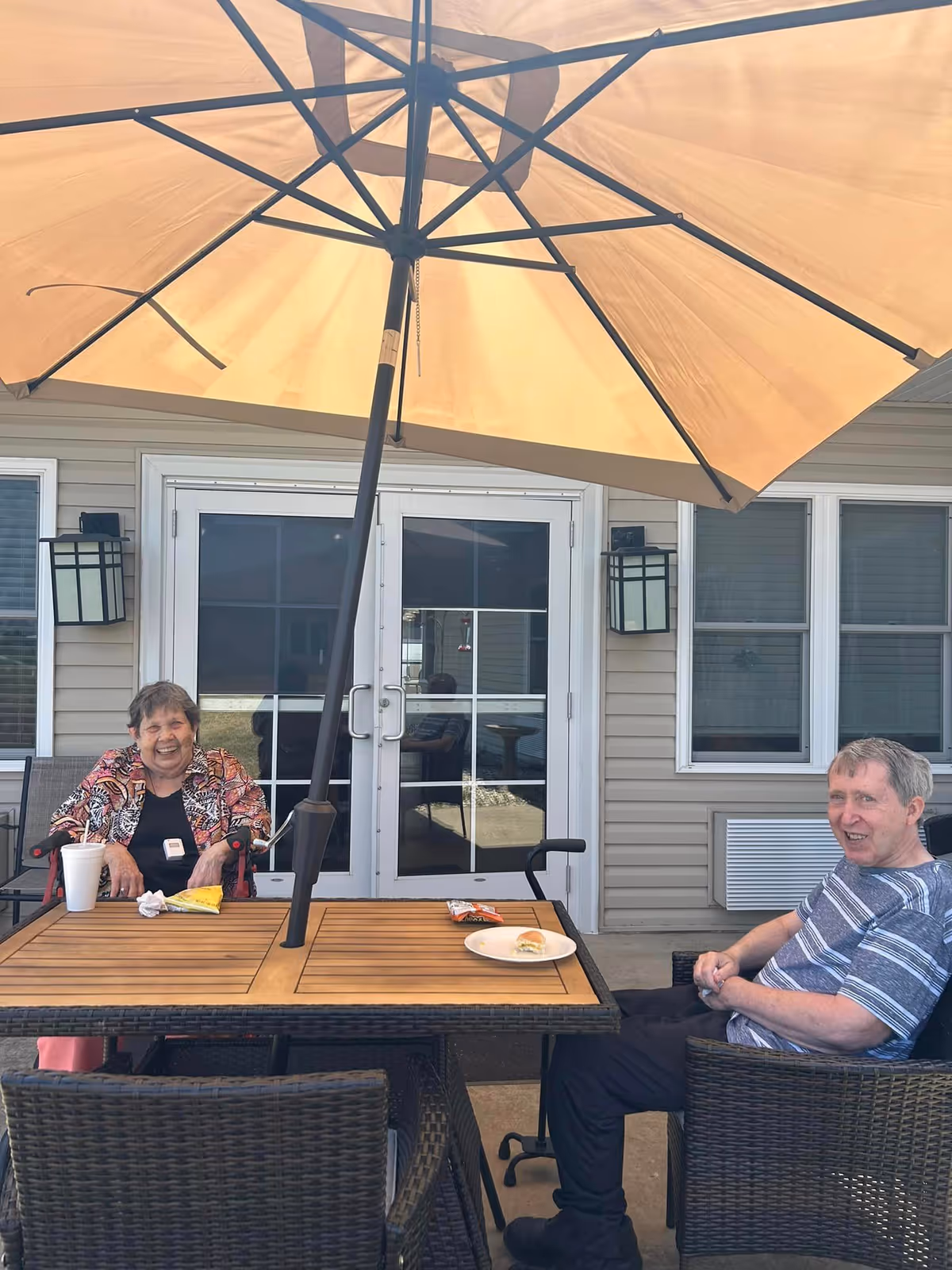 Two elderly people sitting at a wooden outdoor table with a large beige umbrella overhead. They are smiling and appear to be enjoying a meal or snack outside a building with beige siding and white-framed glass doors and windows.