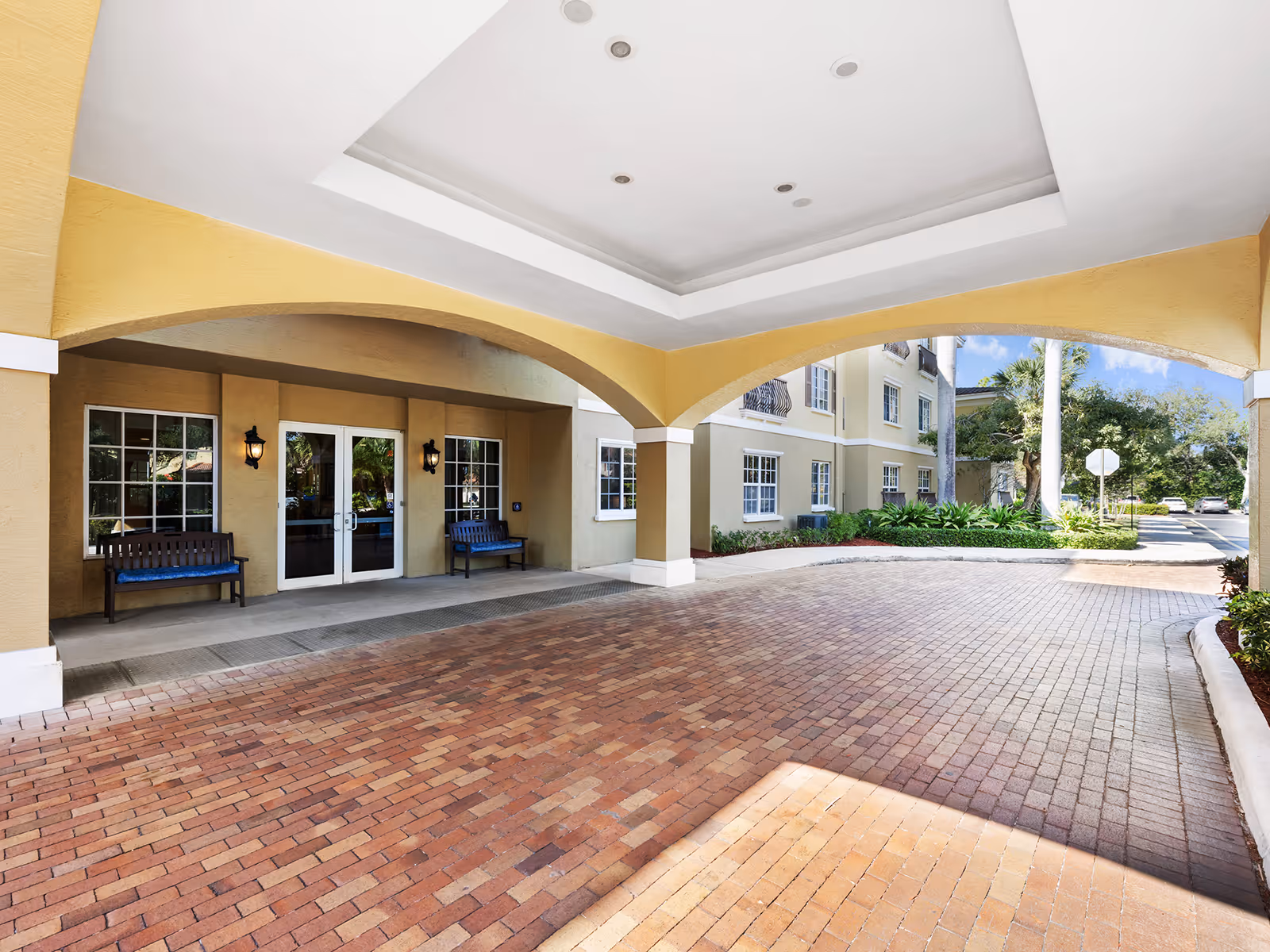 Covered porte-cochere and brick driveway at the front entrance of a beige senior living building with benches and double glass doors.