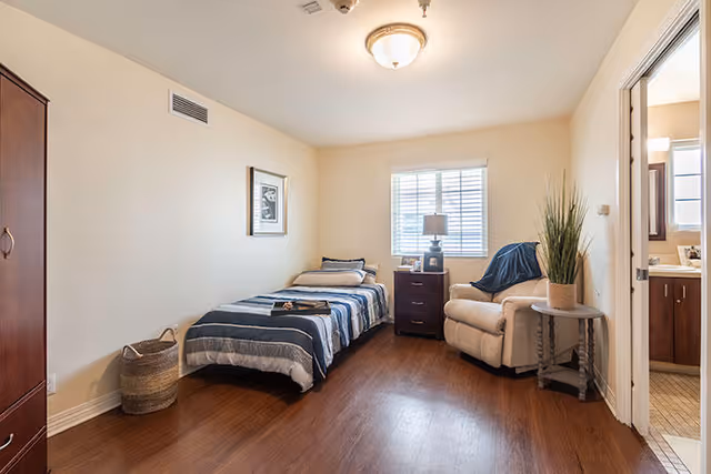 A simple bedroom with a single bed covered in a blue and gray striped blanket, a wooden nightstand with a lamp and picture frames, a beige armchair with a blue throw, a small round side table with a potted plant, a window with white blinds, a framed picture on the wall, and a doorway leading to a bathroom.