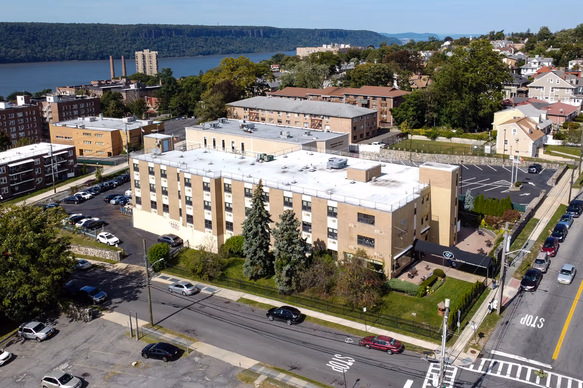 Aerial view of Sans Souci Rehabilitation and Nursing Center, a multi-story beige building with a white roof, surrounded by trees, parked cars, and neighboring residential buildings. A river and forested hills are visible in the background under a clear blue sky.