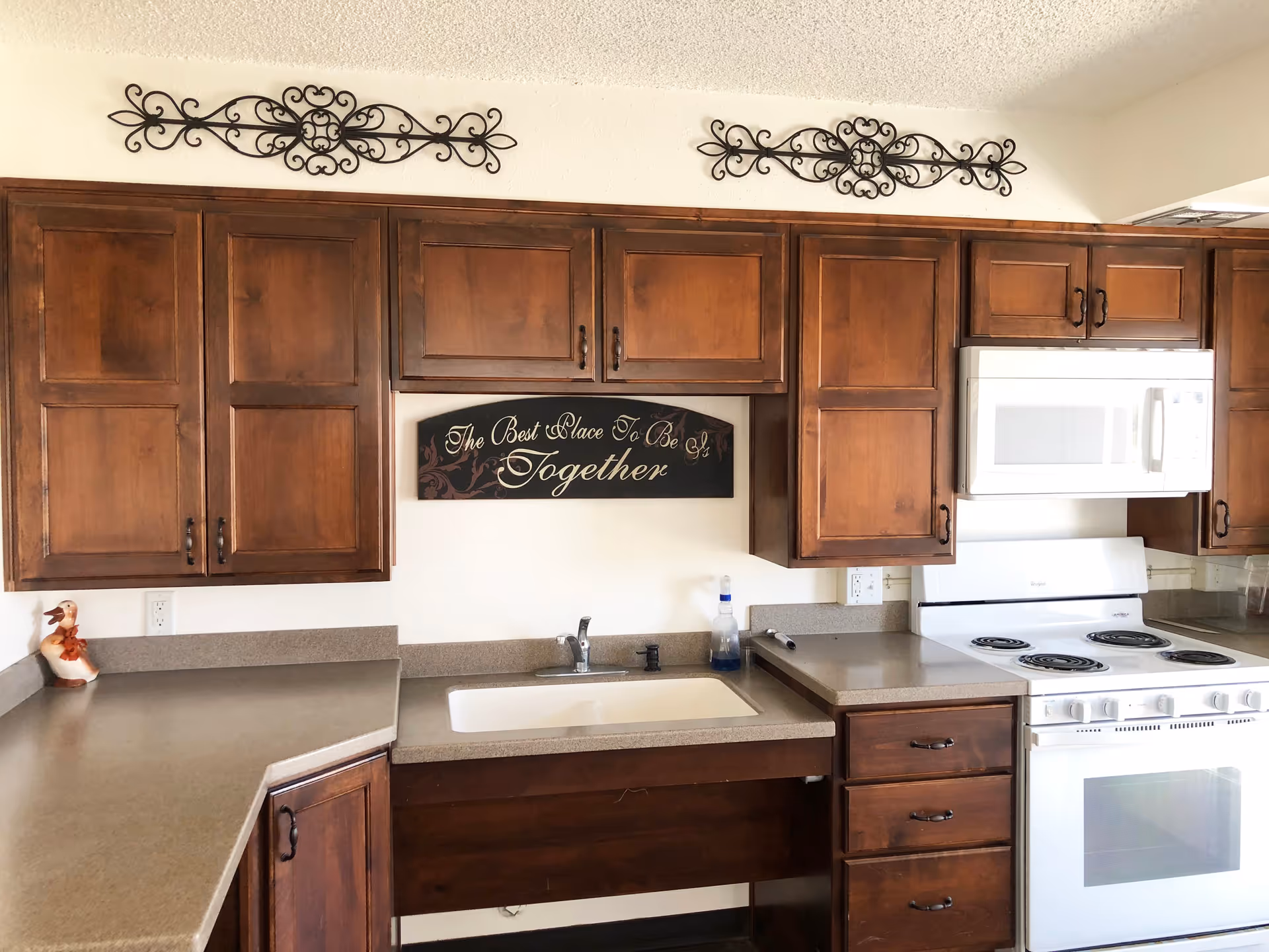 A kitchen with wooden cabinets, a white sink, a white stove with an oven, and a white microwave above the stove. There is a decorative sign on the wall that reads 'The Best Place To Be Is Together' and two ornamental metal wall hangings above the cabinets. A small duck figurine is placed on the left countertop.