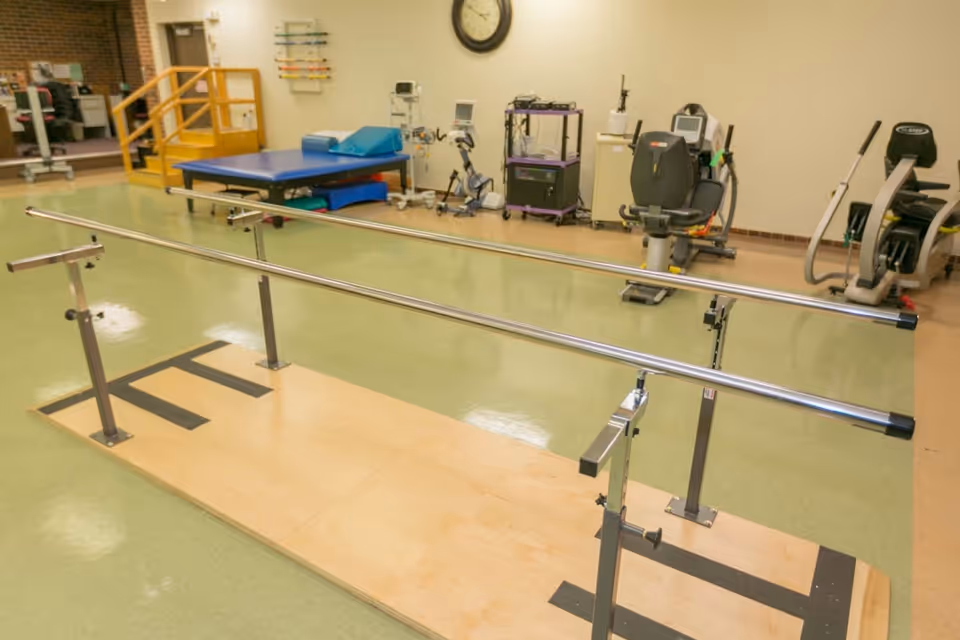 Physical therapy room with parallel bars on a wooden platform and exercise equipment in the background.