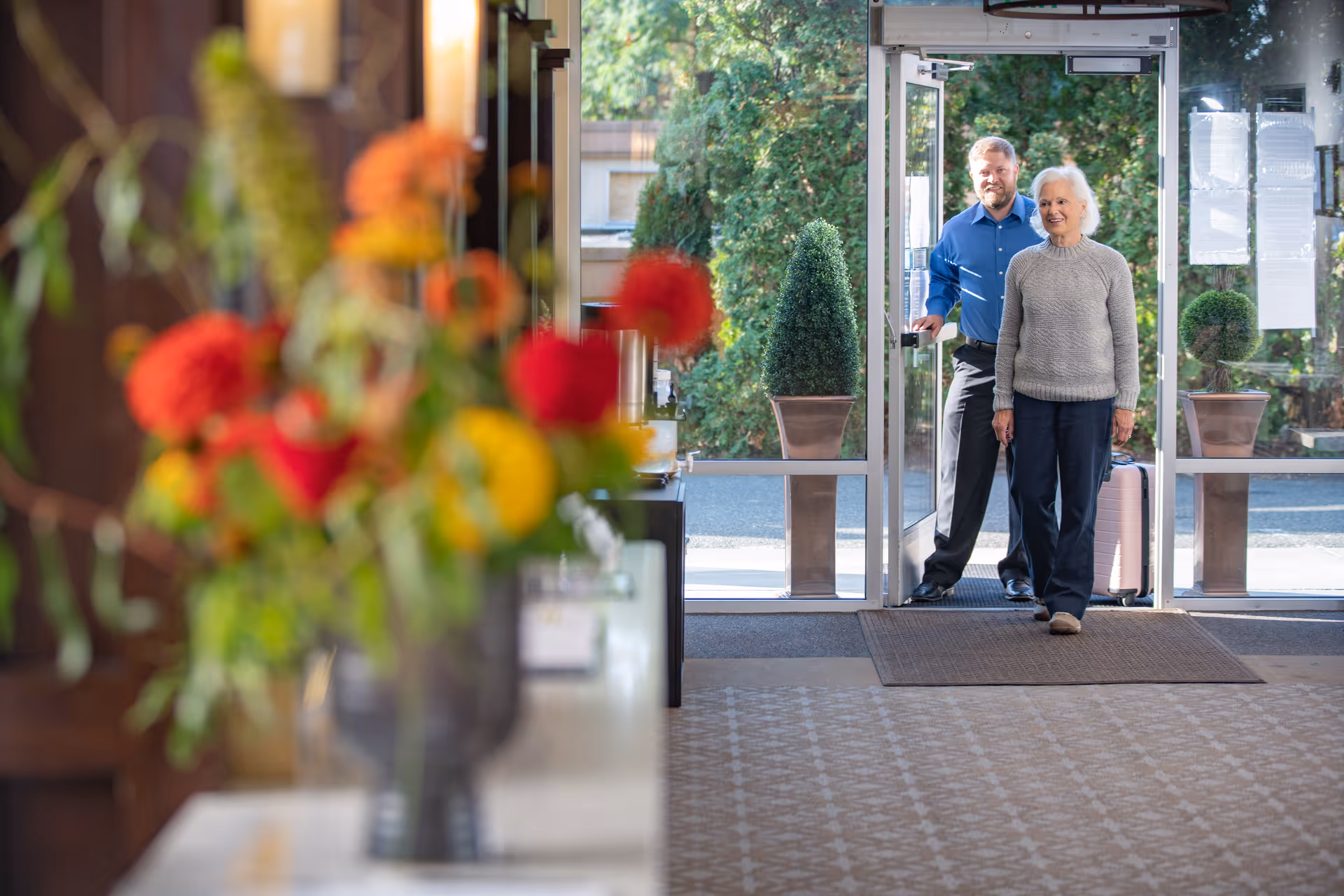 An elderly woman walking inside a building with a man holding the door open behind her. The man is pulling a suitcase. There are large glass doors and potted plants outside. In the foreground, there is a blurred bouquet of colorful flowers on a table.
