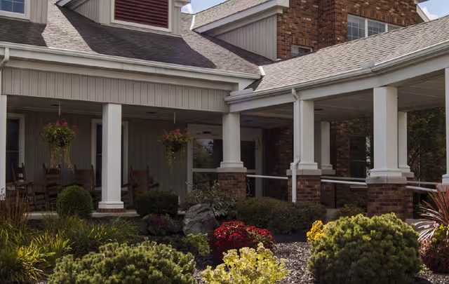 Exterior view of Otterbein Green Hills SeniorLife Community showing a covered entrance with white columns, brick and siding walls, hanging flower baskets, and landscaped garden with various shrubs and flowers.