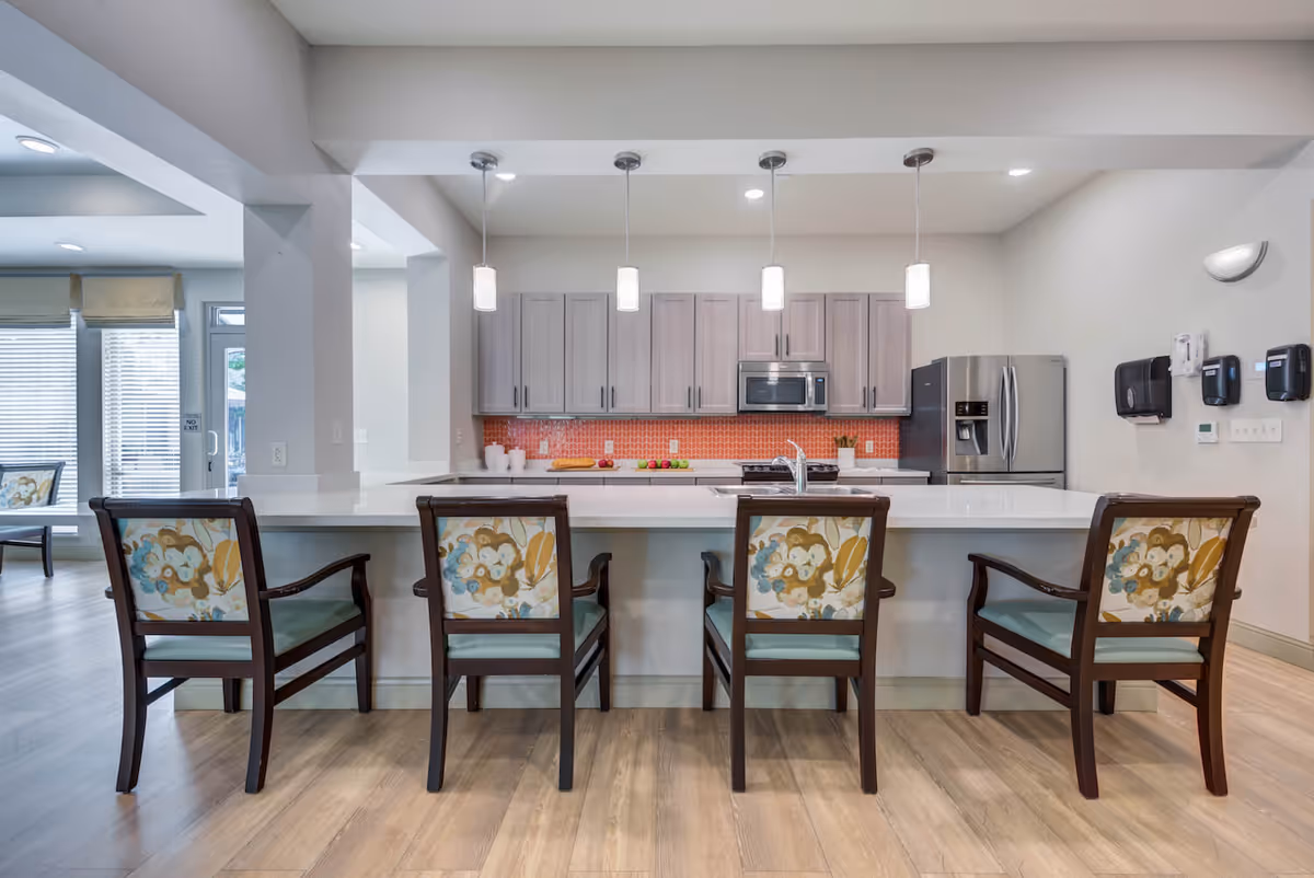 Modern kitchen area with a large white island countertop and four wooden chairs with floral patterned backs and light blue seats. The kitchen features light gray cabinets, a stainless steel microwave, and refrigerator. Four pendant lights hang above the island. The floor is light wood, and there is a red tile backsplash behind the stove.