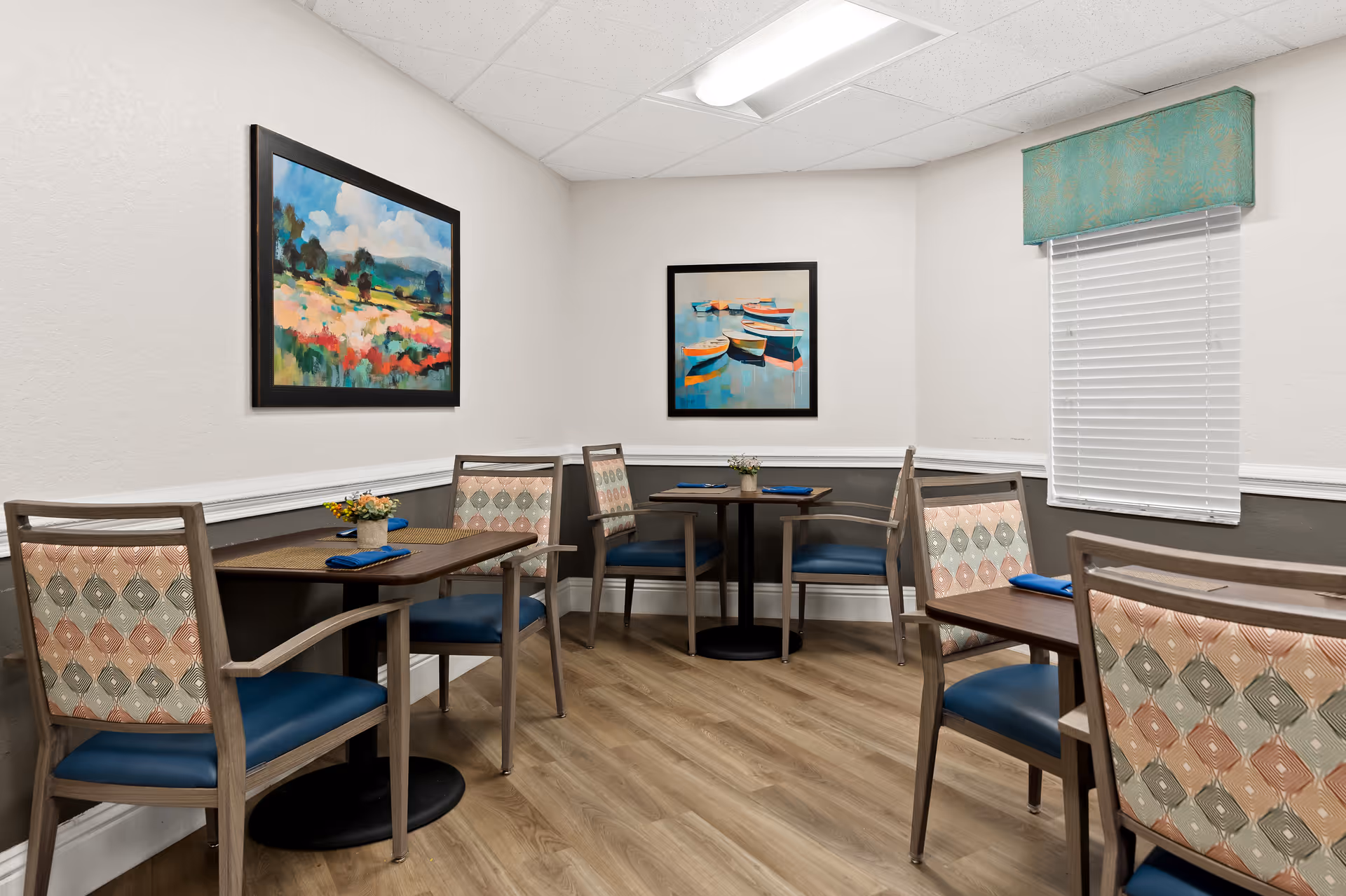 A small dining area with three wooden tables, each set with blue napkins and small flower arrangements. The chairs have patterned upholstery with blue seats. The walls are painted white with a dark gray lower half and decorated with two framed paintings, one depicting a colorful landscape and the other showing boats on water. A window with white blinds and a teal valance is on the right wall. The floor is light wood, and the ceiling has recessed lighting.