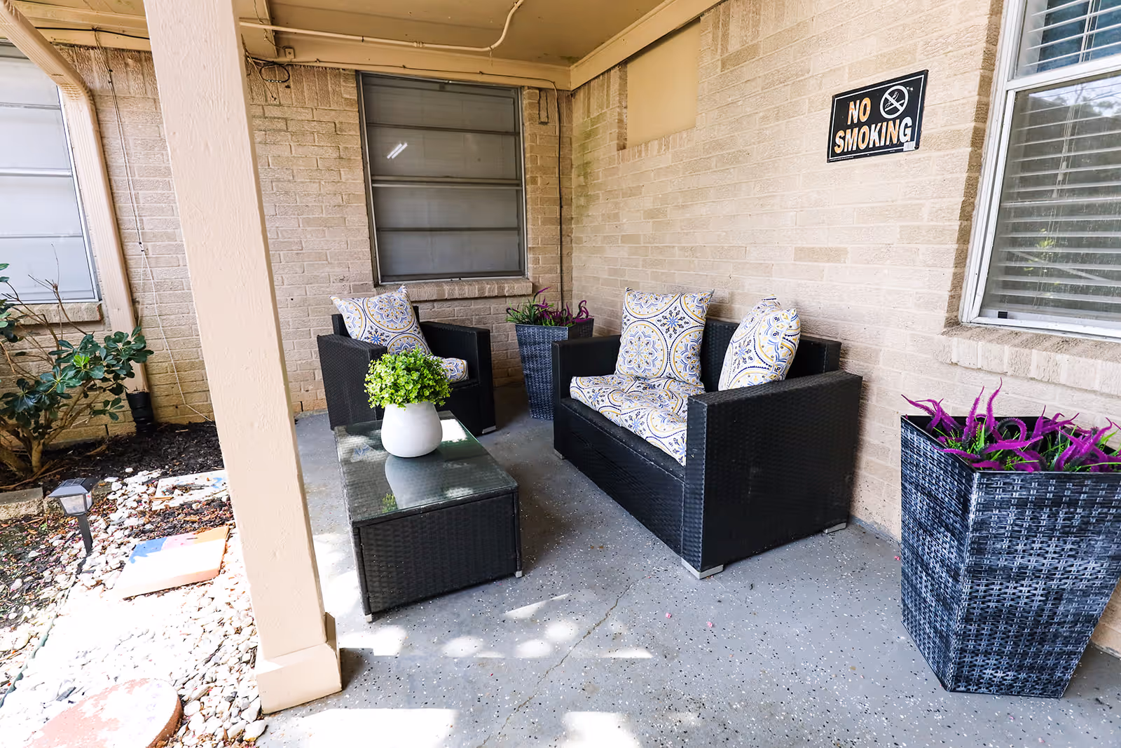 Covered outdoor seating area with wicker sofa and chairs, a coffee table, potted plants, and a 'No Smoking' sign on a brick wall.