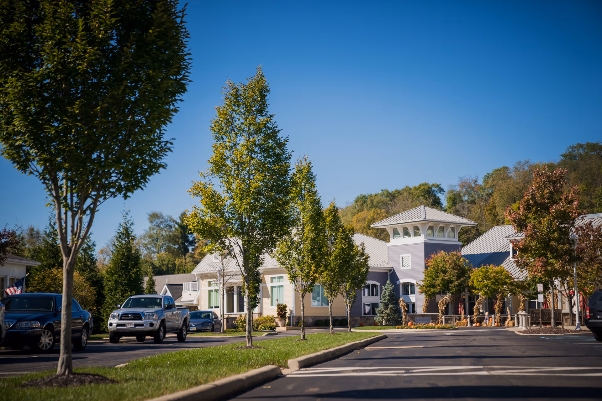 Exterior view of Otterbein Granville SeniorLife Community showing a parking lot with several cars, a row of young trees along the sidewalk, and a building with a distinctive tower and metal roof under a clear blue sky.