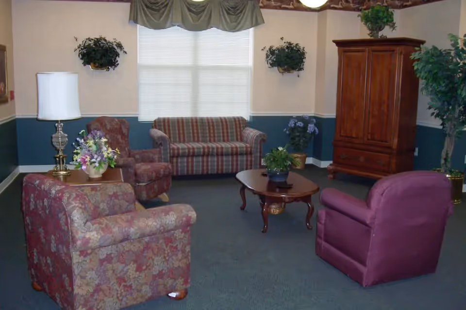 A cozy living room area with a striped sofa, two floral armchairs, and a purple armchair arranged around a wooden coffee table with a potted plant. There is a wooden cabinet in the corner, a table lamp on a side table with a flower arrangement, and several potted plants and wall-mounted greenery. A large window with blinds and a valance lets in natural light.