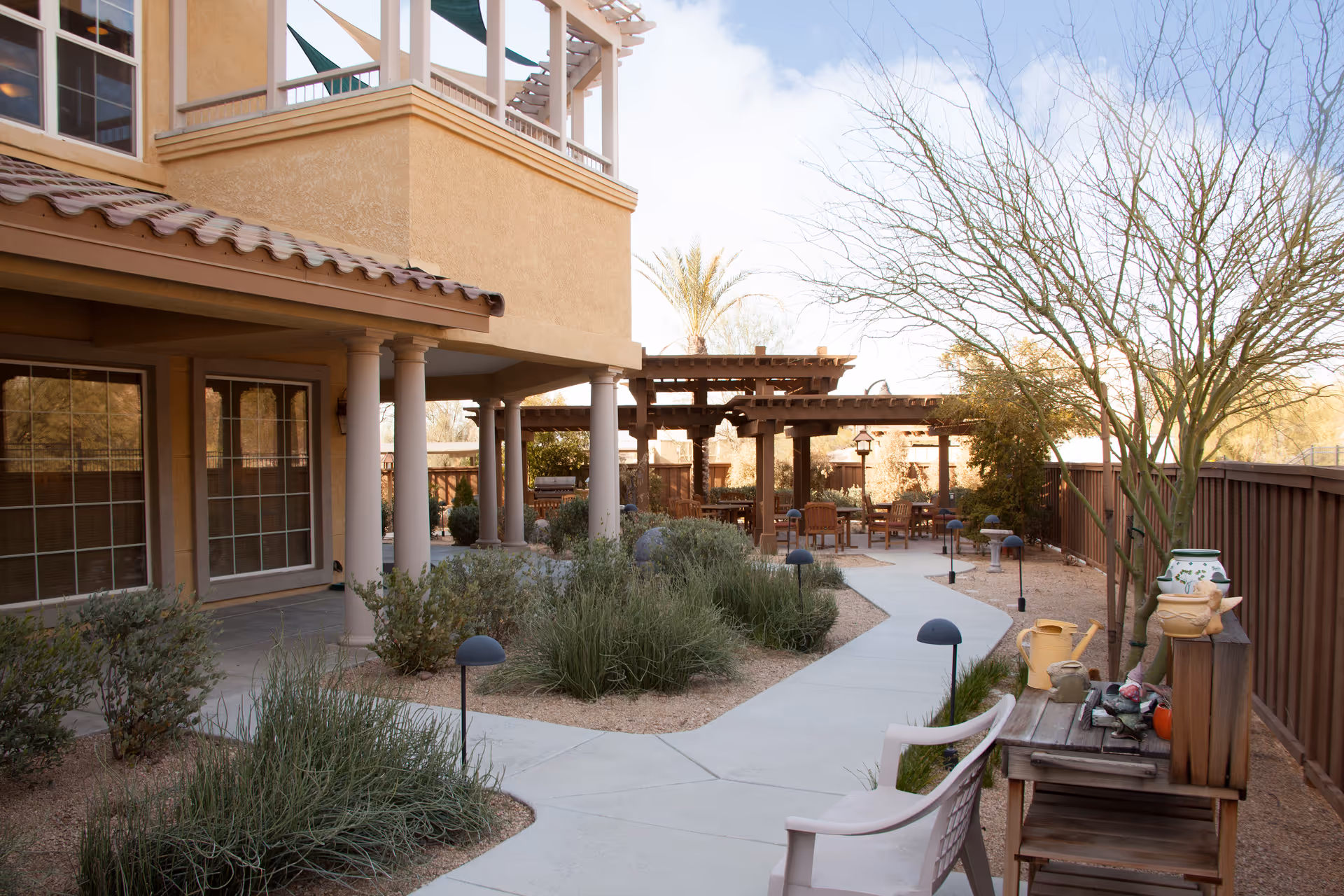Outdoor patio area at Sunrise At River Road featuring a paved walkway, garden beds with shrubs and desert plants, wooden pergolas with seating areas, a wooden table with decorative pots and watering cans, and a beige building with columns and windows in the background.