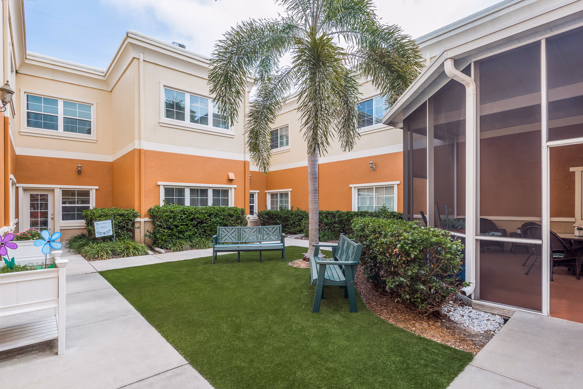 Enclosed courtyard with benches, a palm tree, artificial turf, and a screened patio in front of a beige-and-orange senior living building.