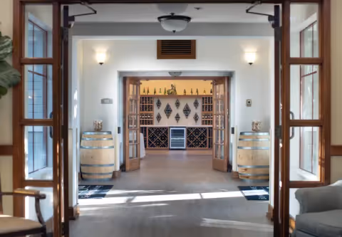 View through an open doorway into a wine storage room with wooden wine racks, wine bottles, and a wine refrigerator. Two wooden barrels are placed on either side of the entrance, and the room is well-lit with wall sconces and ceiling lights. The surrounding area has wooden framed glass doors and a light-colored floor.